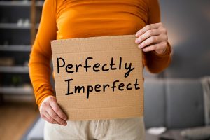 Frau in orangenem Shirt und weißer Hose hält ein Schild hoch auf dem perfekt unvollkommen bzw perfectly imperfect steht