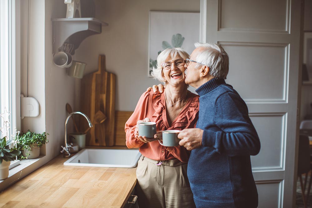 Glückliches älteres Paar in Küche - Mann gibt Frau Kuss auf die Wange
