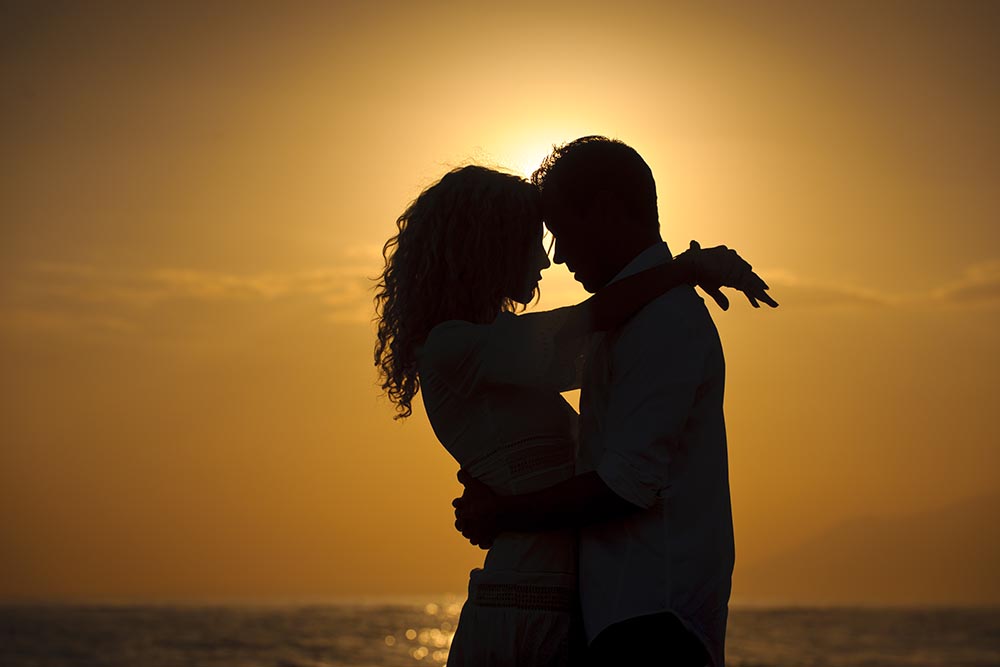 Junges Paar steht eng umschlungen als Silhouetten vor einem orangenen Sonnenuntergang am Meer