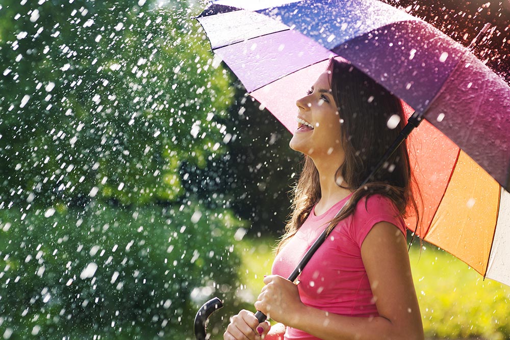 Frau steht lachend mit einem Regenbogen-Regenschirm im Regen und findet Perfektion in Unvollkommenheit