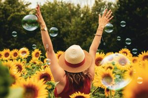 Frau steht mit hochgestreckten Armen und Strohhut in einem Feld aus Sonnenblumen und versprüht Energie und Lebensfreude