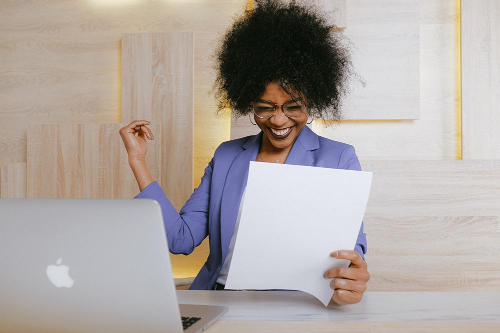 Junge schwarze Frau blickt vor ihrem Laptop breit lächelnd auf ein Blatt Papier und macht mit ihrem Arm eine starke Geste die Erfolg symbolisiert
