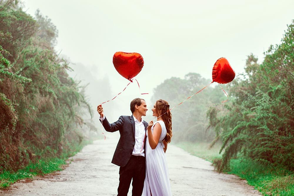 ein junges frisch verheiratetes Paar sieht sich verliebt an und hält Herzluftballons die über ihnen schweben