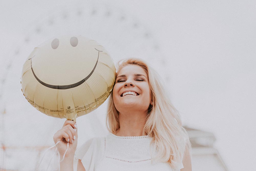 junge schöne blonde Frau steht mit einem Smiley-Ballon vor einem Jahrmarkt und lacht vor Lebensfreude