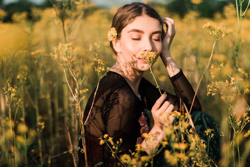junge Frau sitzt unbeschwert in einem Feld aus Blumen und riecht zufrieden mit geschlossenen Augen an einer gelben Blüte