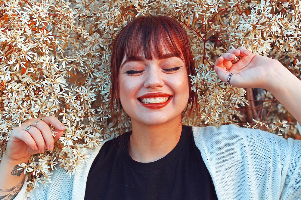 junge rothaarige Frau lächelt mit geschlossenen Augen vor Lebensfreude vor einem Hintergrund aus weißen Blüten
