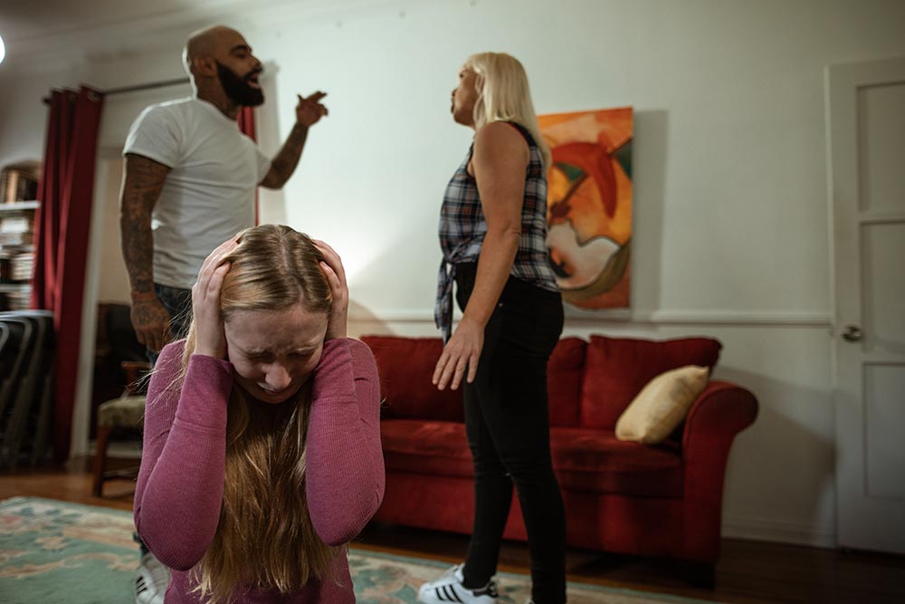 Teenager-Mädchen hält sich im Vordergrund weinend die Ohren zu während ihre Eltern im Hintergrund streiten