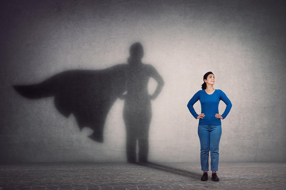 Brünette junge Frau steht selbstbewusst da während ihr Schatten hinter ihr an der Wand sie mit Superhelden-Umhang zeigt