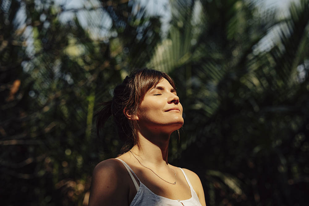 Junge Frau steht während Kontaktsperre glücklich und entspannt mit geschlossenen Augen im Grünen und hält ihr Gesicht in die Sonne