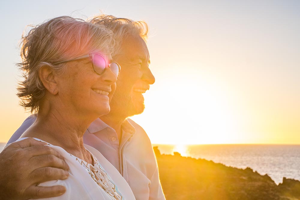 Seniorenpaar sitzt zufrieden lächelnd während des Sonnenuntergangs am Strand während er seinen Arm um ihre Schultern gelegt hat