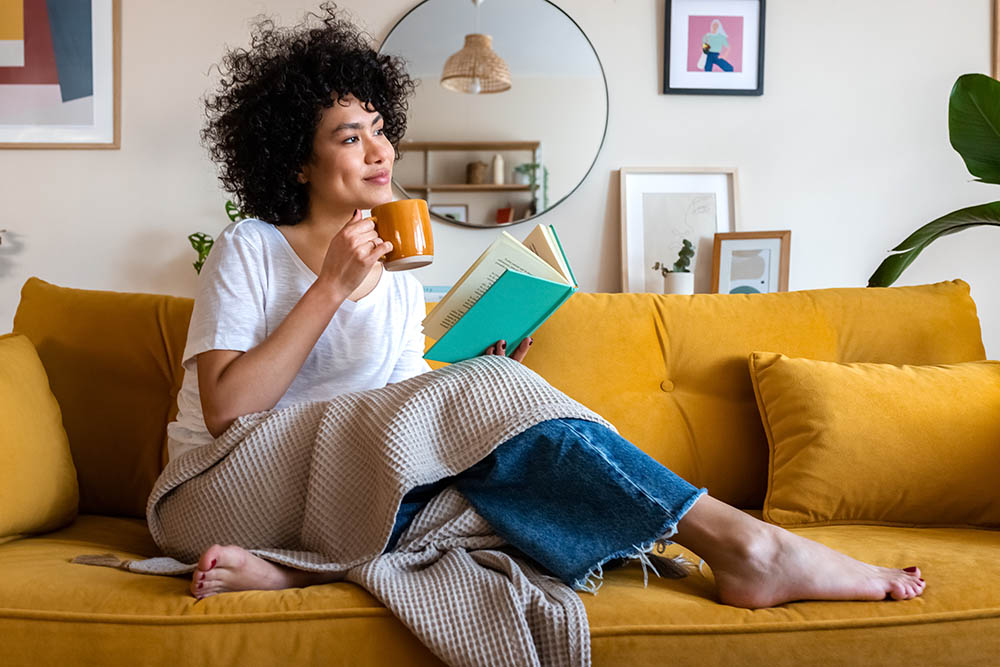 Junge Frau sitzt während Kontaktsperre glücklich mit einem Buch einer Decke und einer Tasse Tee auf der Couch
