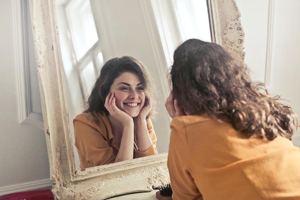 junge brünette Frau betrachtet sich lächelnd und selbstbewusst im Spiegel