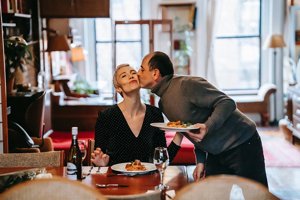 Mann serviert Frau Essen dass er für sie gekocht hat und küsst sie auf die Wange