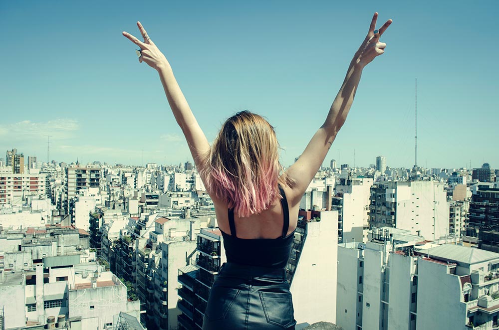 junge blonde Frau schaut selbstbewusst über eine Stadt und streckt die Hände energetisch gen Himmel