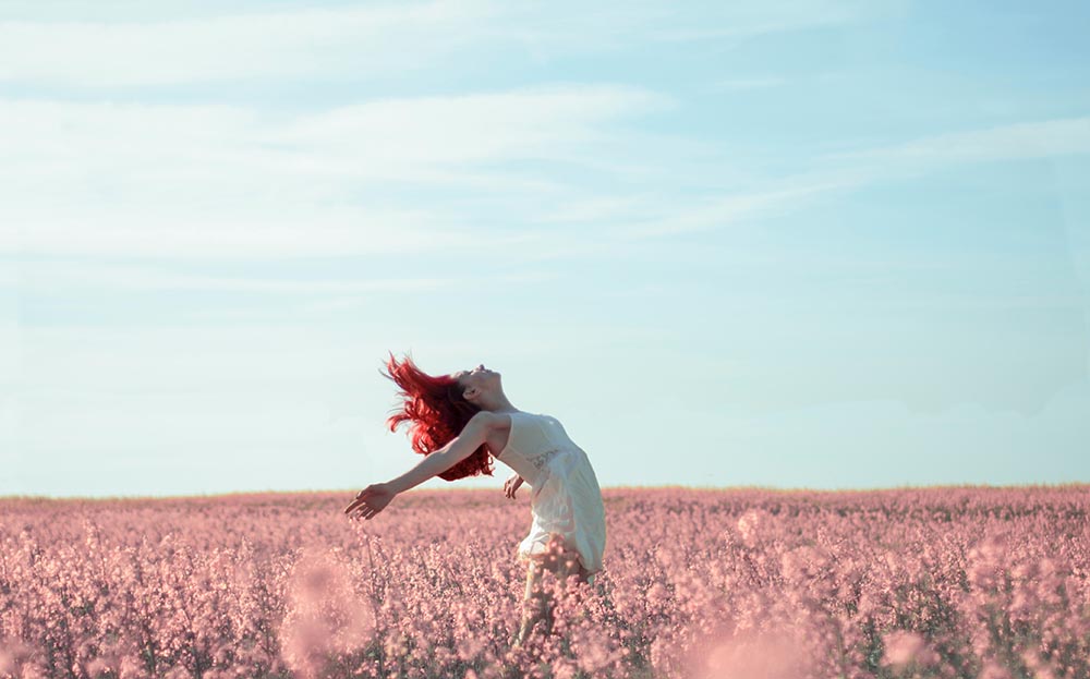junge Frau mit knallig roten Haaren streckt sich selbstbewusst und frei in einem Feld aus rosa Blumen und ist offen für neue Abenteuer
