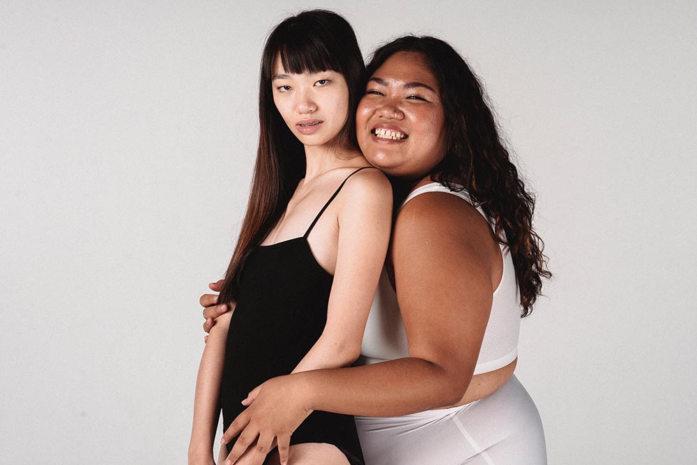 eine schlanke asiatische und rundliche schwarze Frau lachen in Unterwäsche lachen im Rahmen der Body Positivity Bewegung selbstbewusst und glücklich in die Kamera