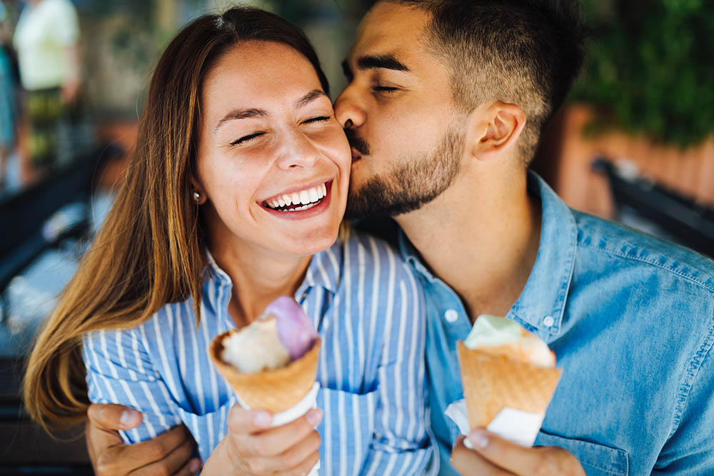 junger Mann küsst junge lachende Frau auf die Wange während beide Eistüten halten
