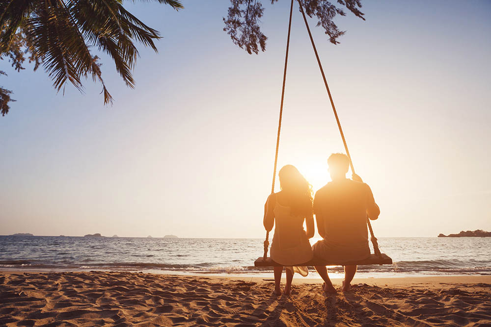 junges Paar sitzt gemeinsam auf einer Schaukel am Strand und blickt glücklich in den Sonnenuntergang