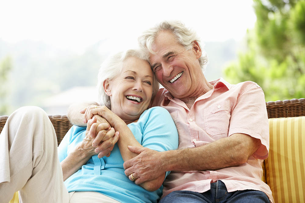 reife Frau und reifer Mann sitzen als älteres Paar glücklich lachend und sich umarmend auf einer Bank im Grünen