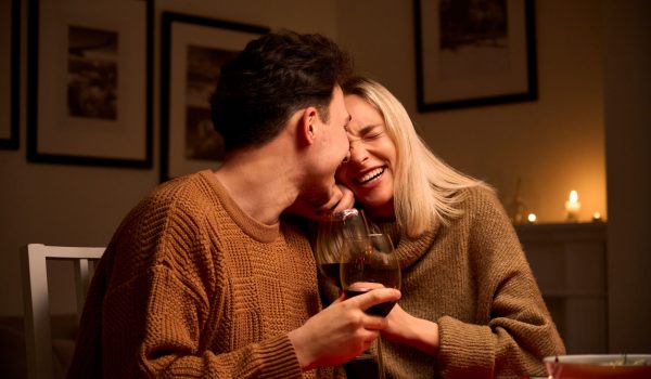 Happy young couple in love hugging, laughing, drinking wine, enjoying talking, having fun together celebrating Valentines day dining at home, having romantic dinner date with candles sitting at table.